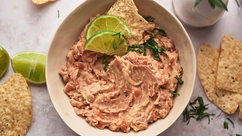 A bowl of refried beans with lime wedges and tortilla chips.