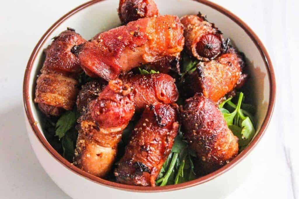 Bacon wrapped sausages in a bowl on a white table.