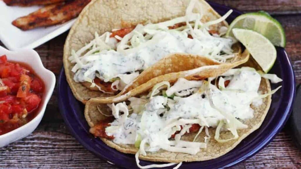 Two fish tacos on a plate with limes and tomatoes.