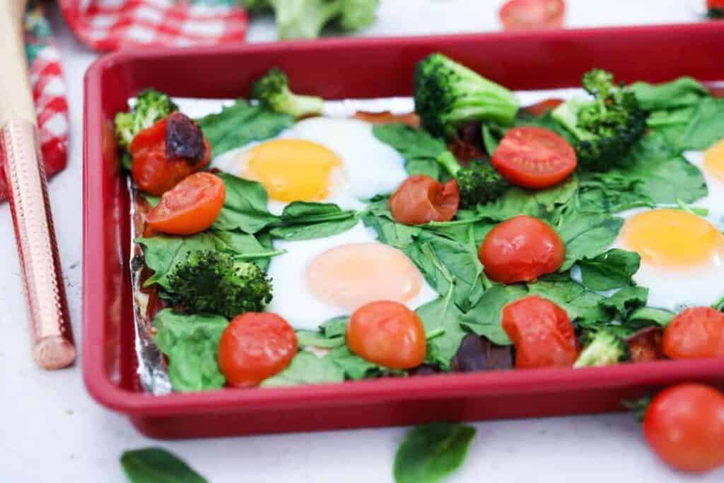 A red baking dish with eggs, tomatoes and broccoli.