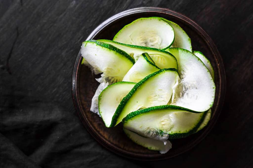 Sliced zucchini in a wooden bowl on a dark background.