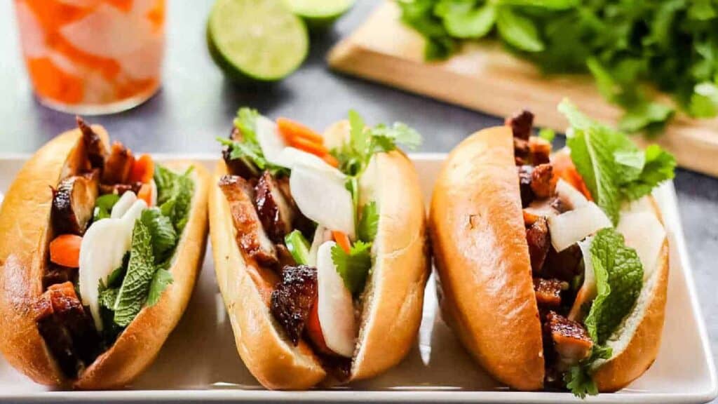3 pork belly banh mi on a white platter with limes and pickled veg in the background.