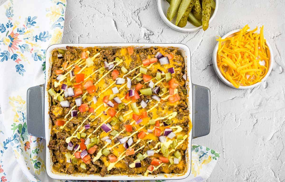 A top-down shot of Cheeseburger Casserole with a bowl of pickles and cheese nearby.