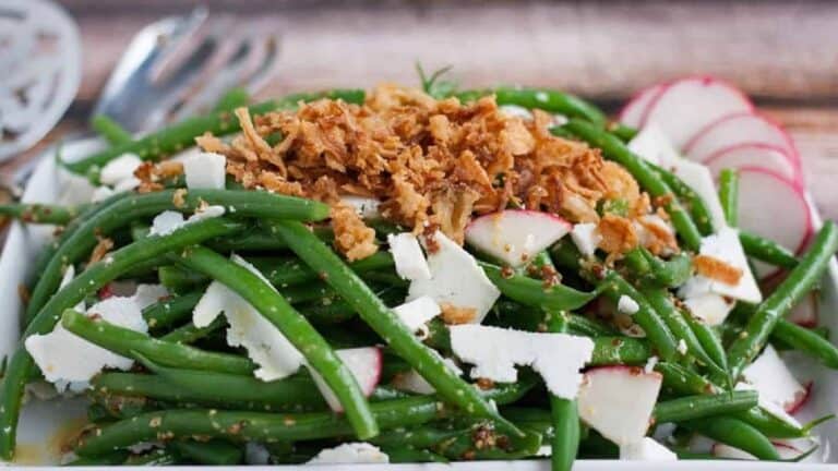 Low angle shot of green bean salad with fried onions and dry ricotta on a white plate.