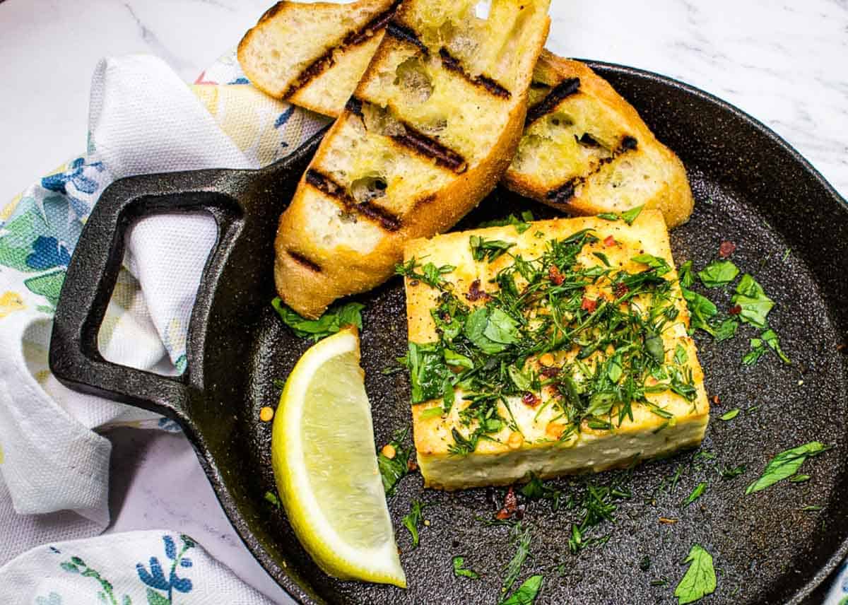 Grilled Feta with Herbs in a black serving dish with toasted bread and a lemon wedge.