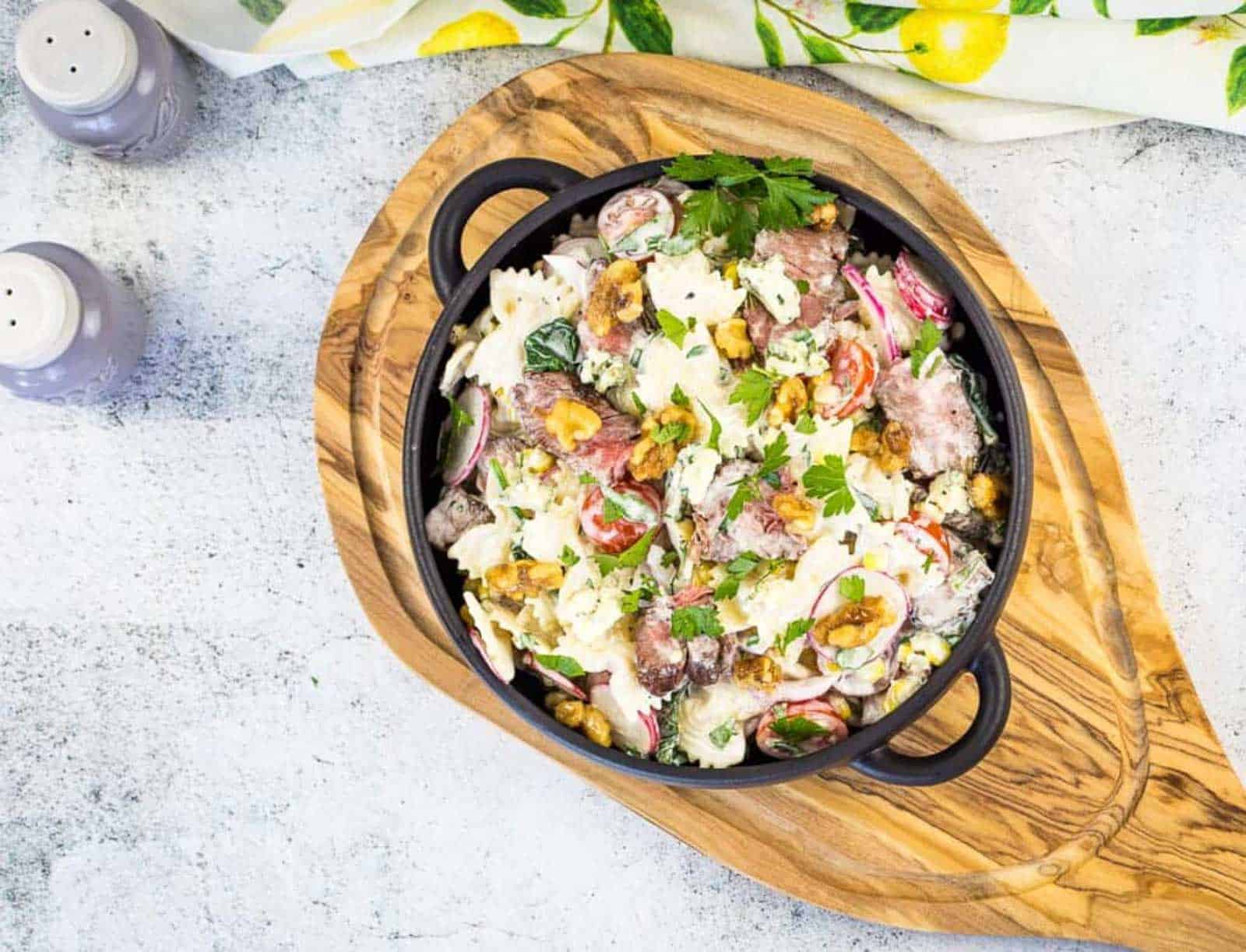 A black bowl filled with a pasta salad containing farfalle, steak slices, radishes, cherry tomatoes, lettuce, nuts, and crumbled blue cheese, garnished with parsley, on a wooden board next to a spoon.