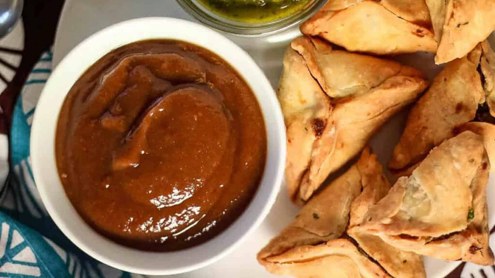 Overhead shot of a bowl of tamarind date chutney with samosas and cilantro mint chutney.
