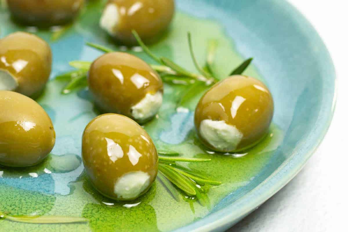 a closeup of blue cheese inside of green olives smothered in olive oil and surrounded by fresh rosemary on a blue plate.