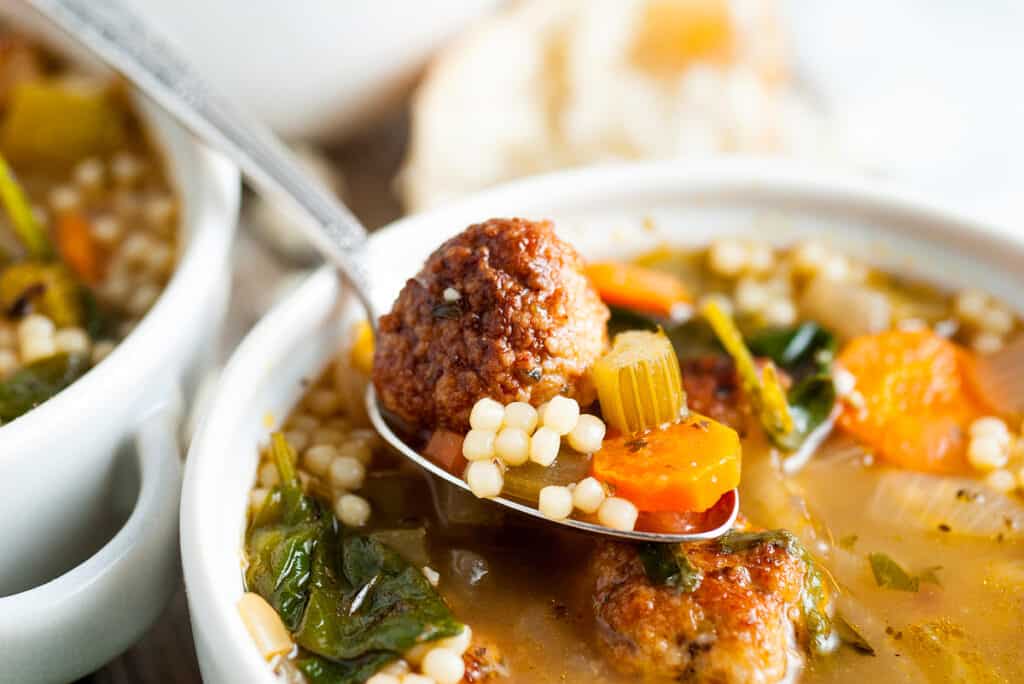 A spoon scooping out from a bowl of Italian wedding soup with meatballs.