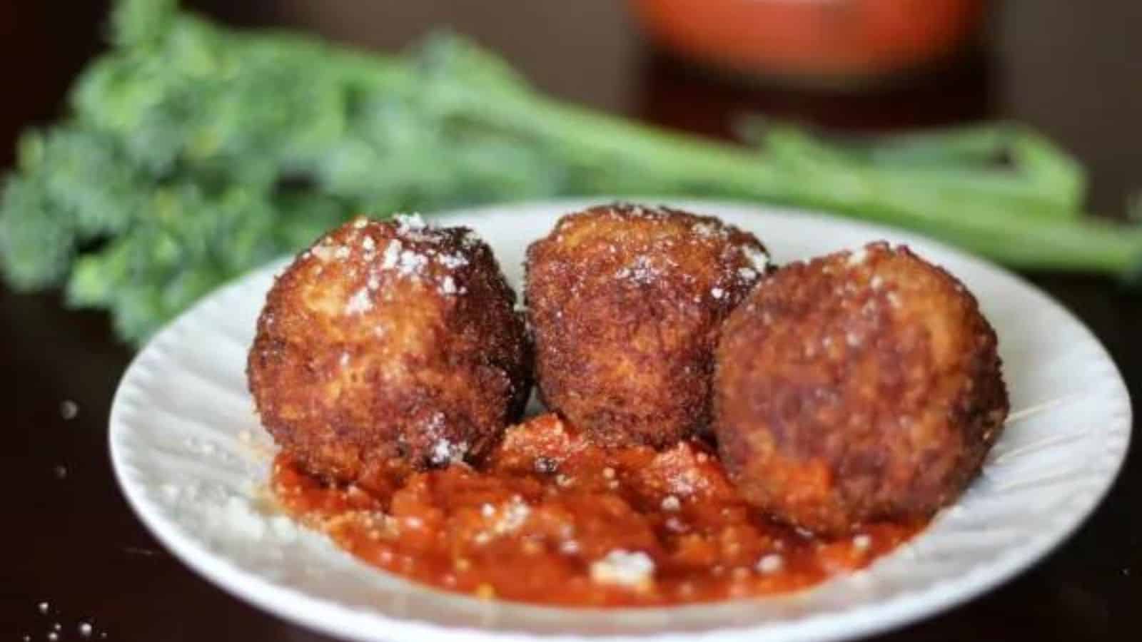 Plate of arancini with marinara and broccolini blurred in background.