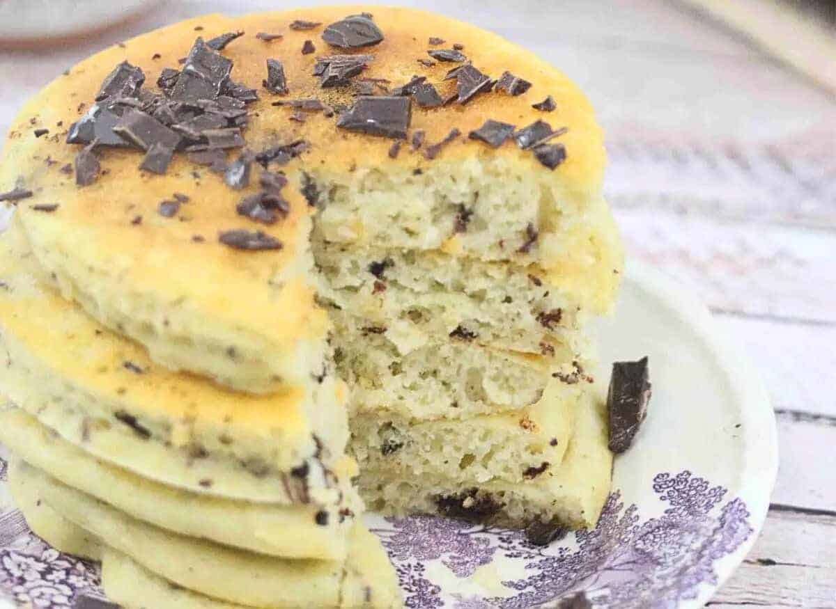 Protein pancakes with chocolate chips stacked on a white and purple platye with a slice cut out of the stack.