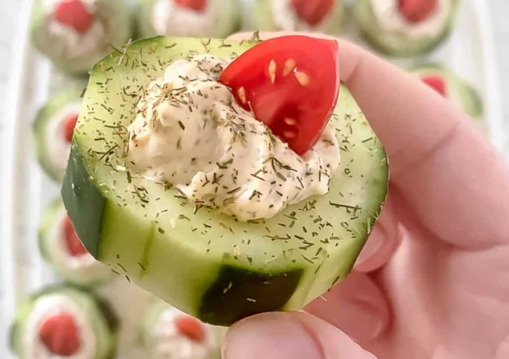 A person holding a cucumber with a dip and tomato on it.