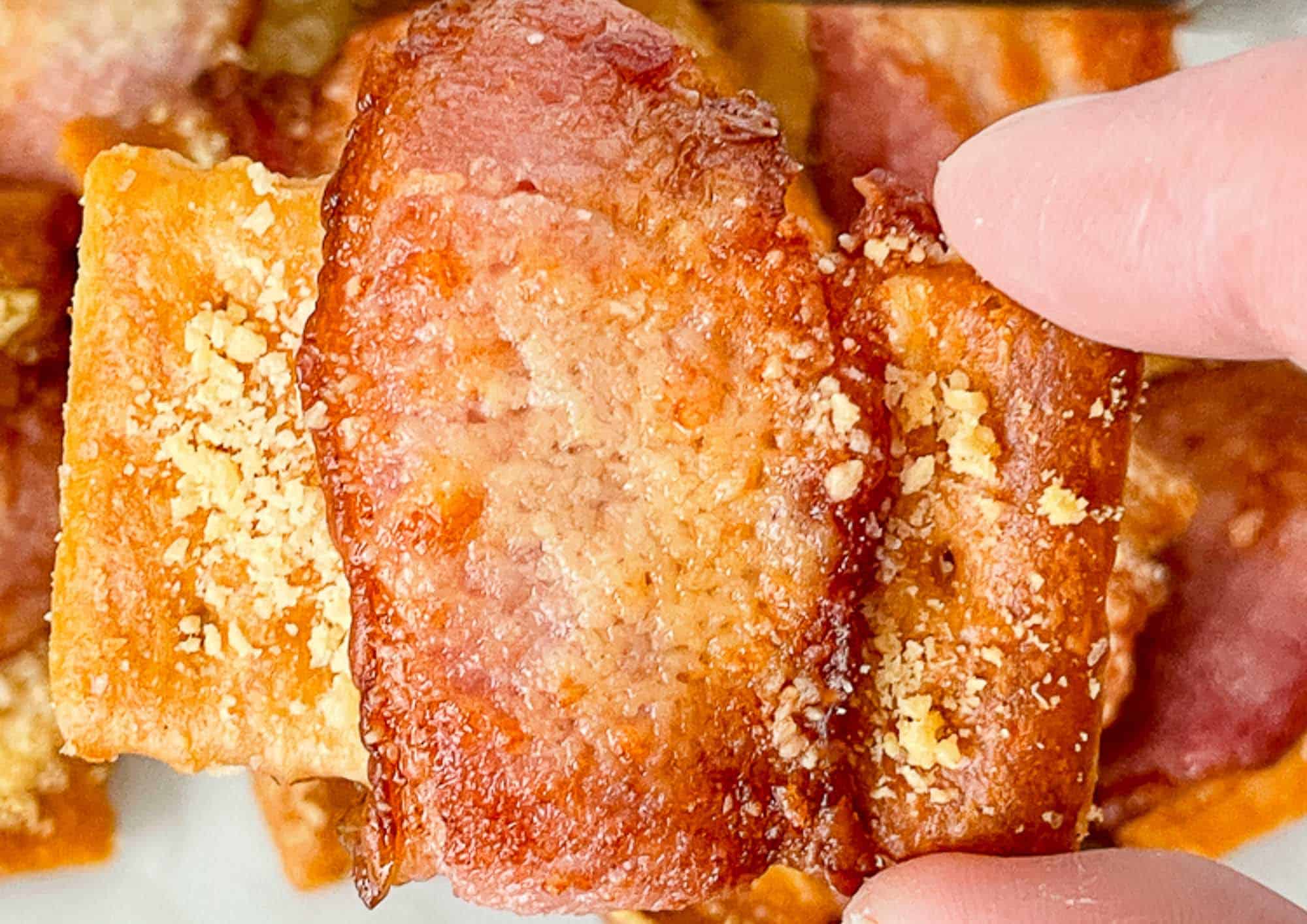 Fingers holding a bacon wrapped crackers.