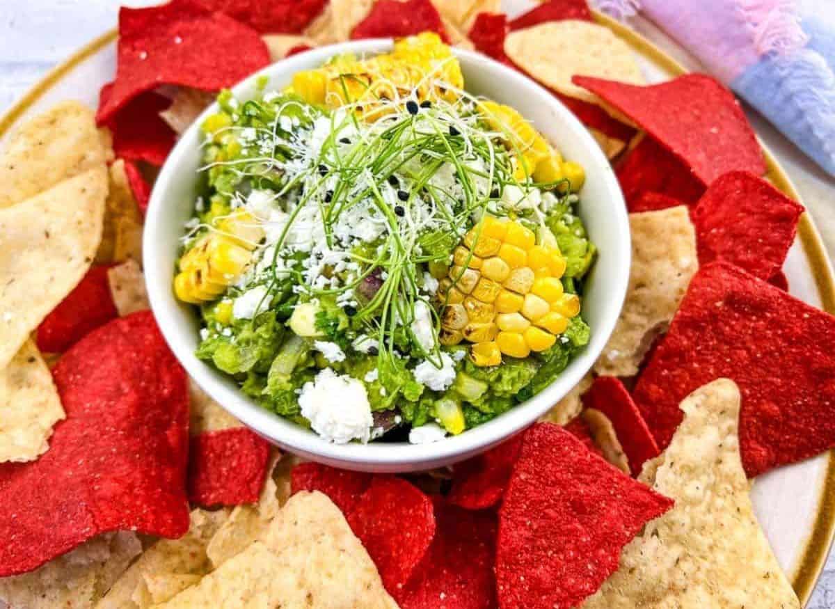 A bowl of Grilled Corn Guacamole surrounded by white and red tortilla chips.