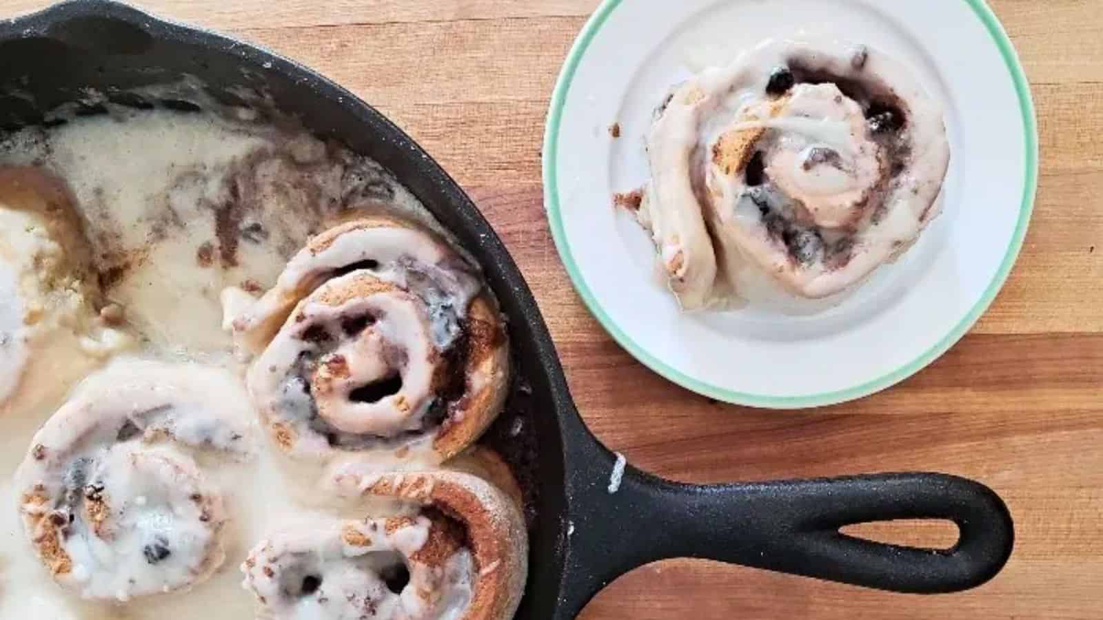 Image shows an overhead shot of Cinnamon rolls in a skillet on a wooden table with one cinnamon roll on a plate.
