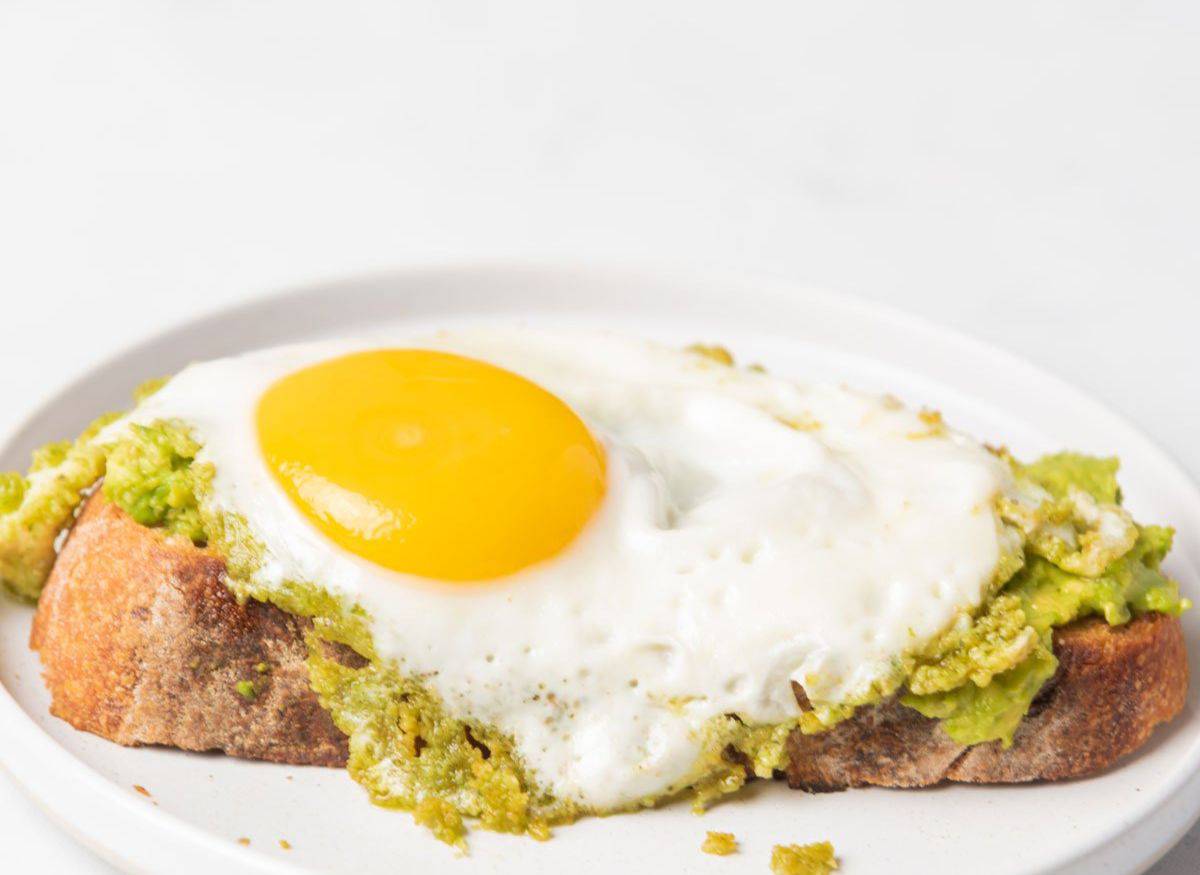 A cooked sunnyside up egg with pesto on top of a slice of sourdough bread covered in mashed avocado on a small round white plate.