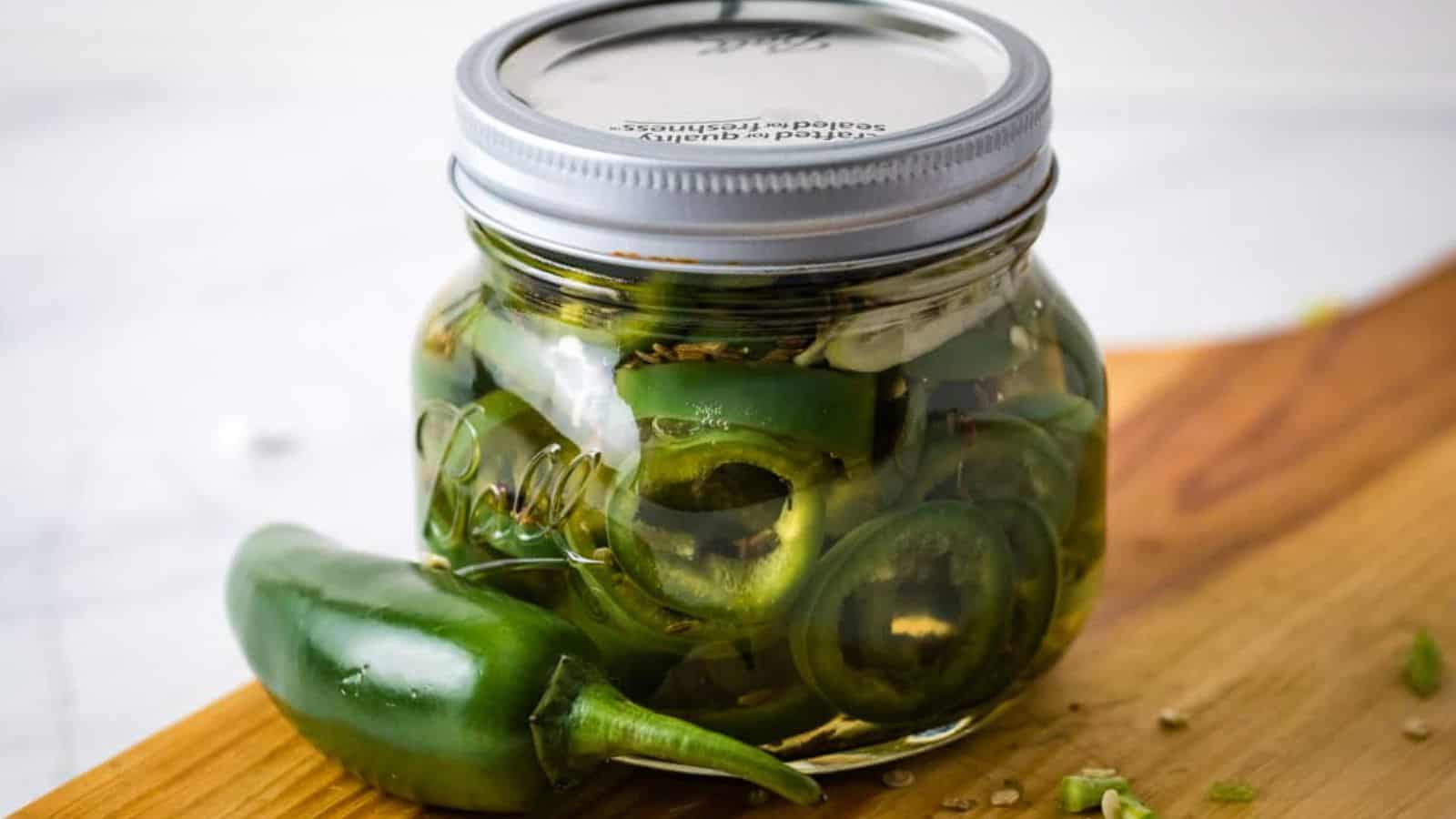 Jar of pickled jalapenos on a cutting board with a pepper next to it.