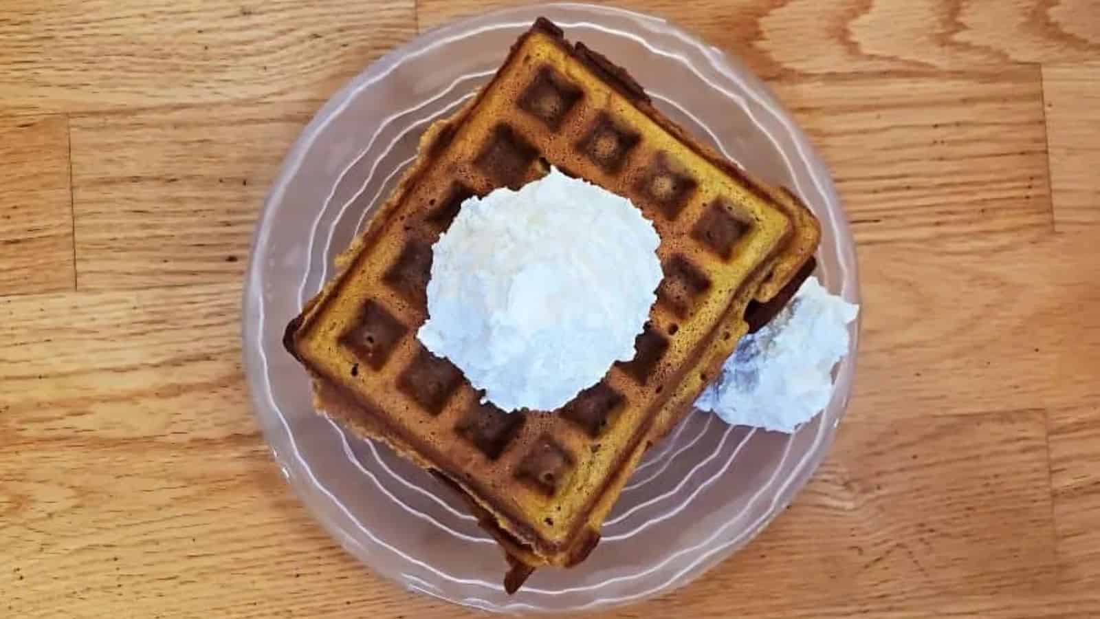 Image shows an overhead shot of Pumpkin Waffles with whipped cream on a glass plate.