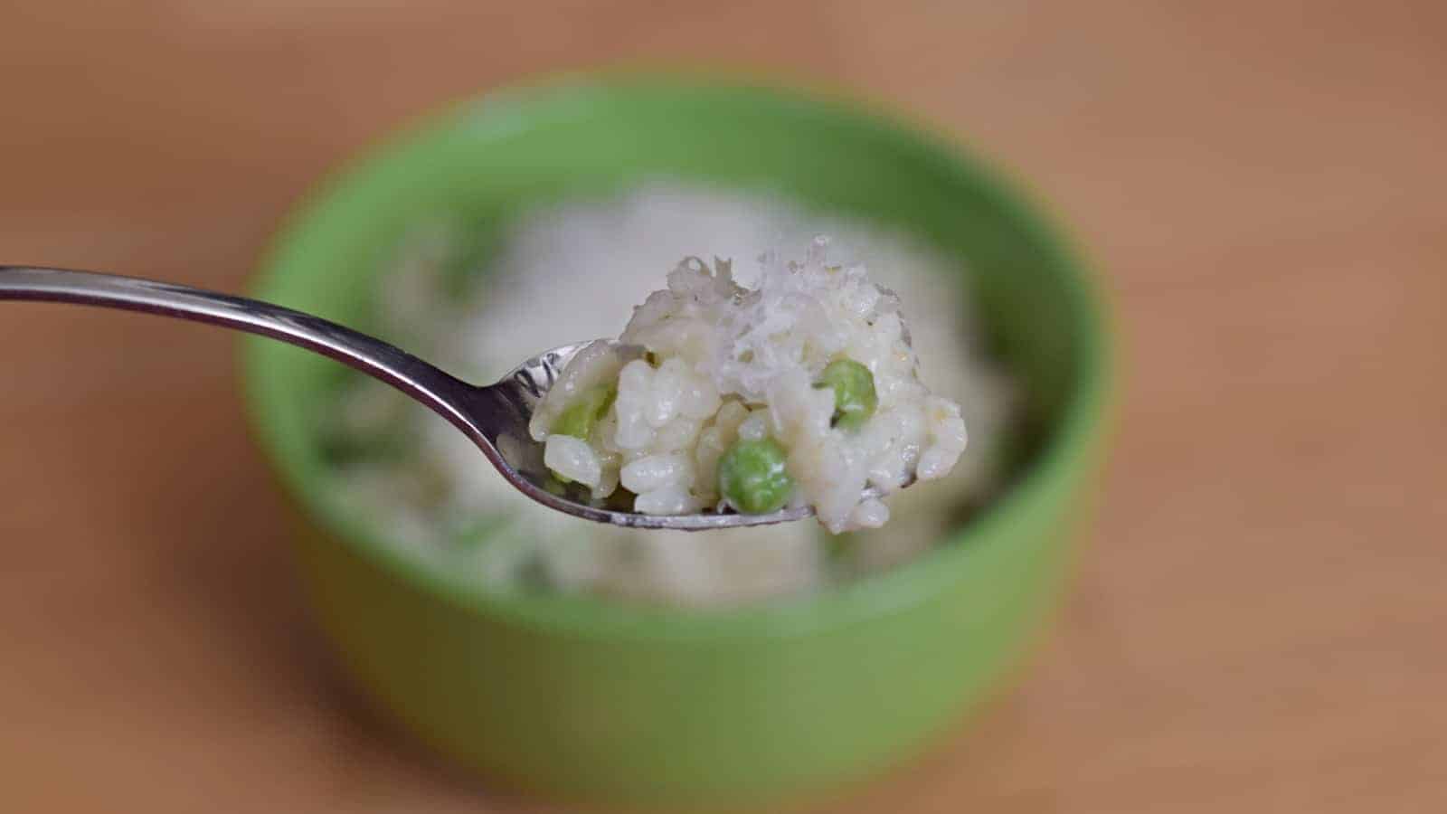 Image shows a Spoonful of risotto with peas and lemon with the rest of the bowl in the background.