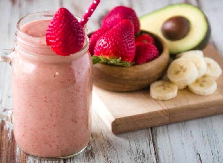 Mug of strawberry smoothie with avocado and banana topped with a strawberry and a red straw.