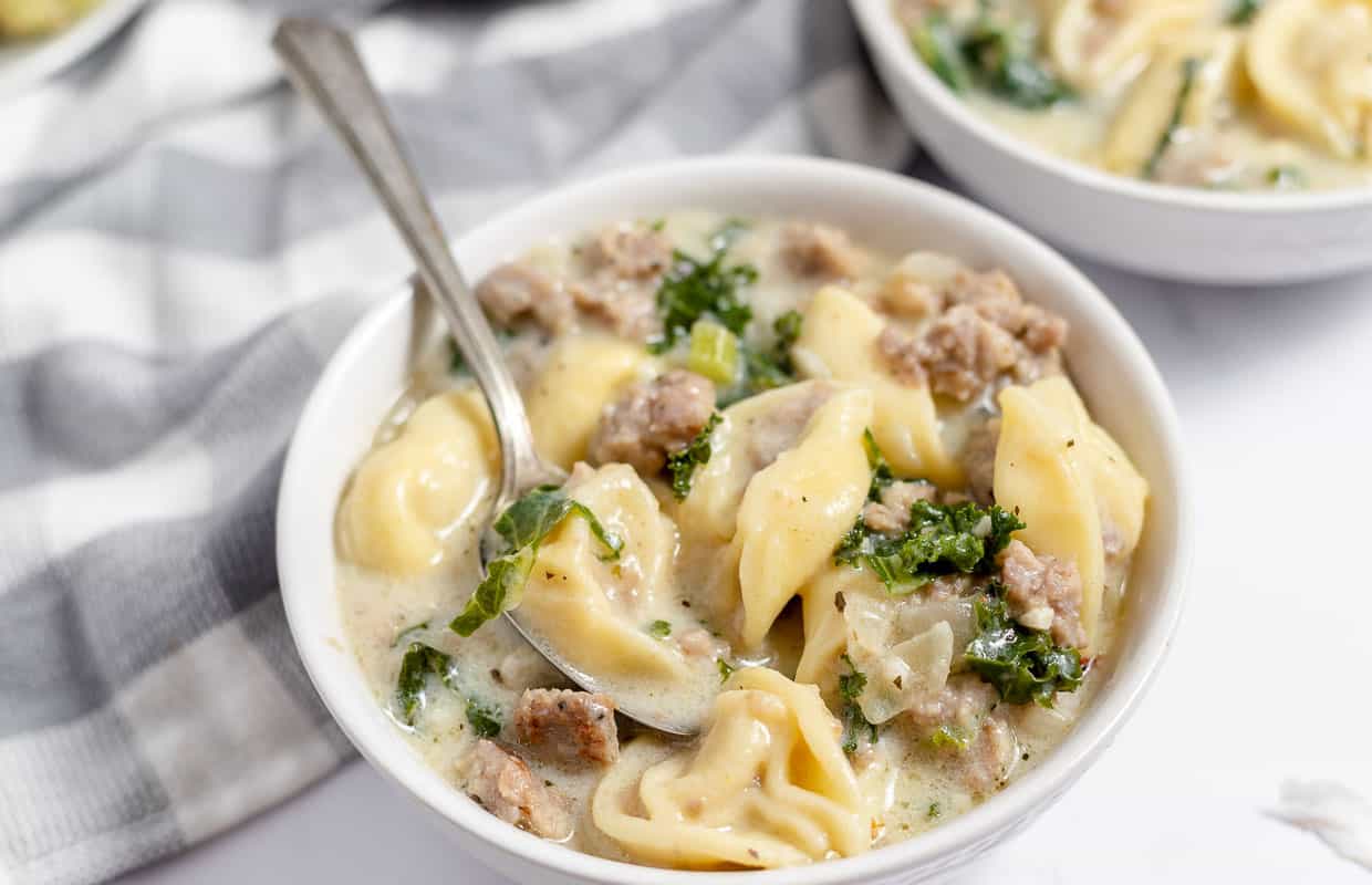 A spoon holidng sausage and tortellini soup over a bowl of soup.