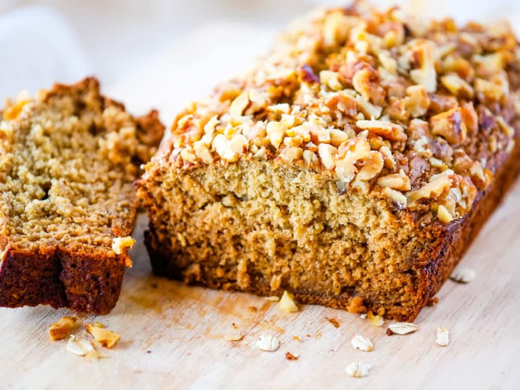 Loaf of banana bread sliced to show the insides with a piece laying on a wooden cutting board.