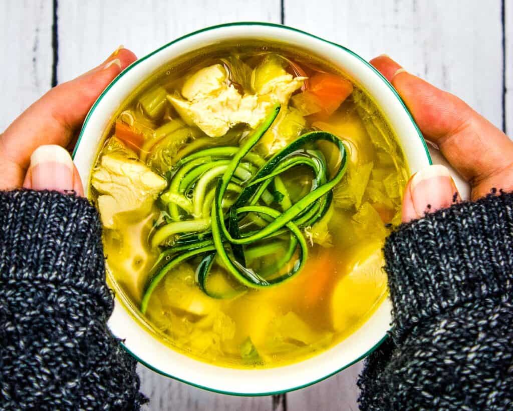 A person holding a bowl of chicken and zucchini soup, one of the soups that taste better.