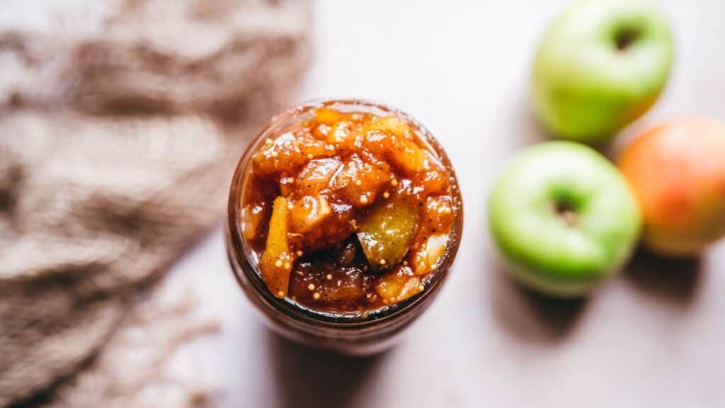 A jar of apple chutney, a condiment, on a table next to apples.