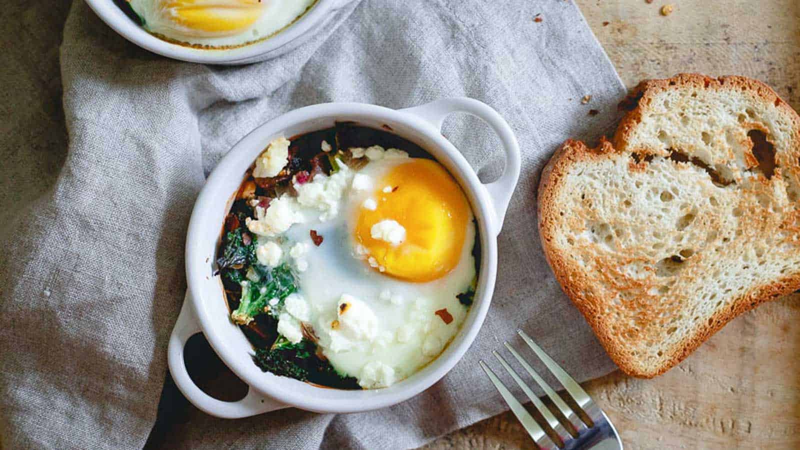 Two bowls of kale feta egg bake with spinach and bread on a table.