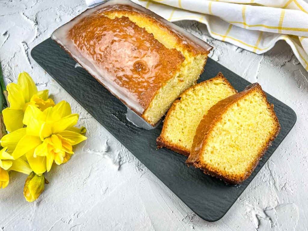 A slice of lemon pound loaf on a plate with daffodils, a sweet treat to love.