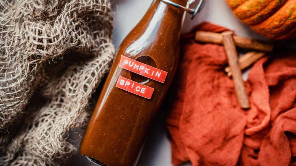 Description (modified): A bottle of pumpkin sauce next to a pile of pumpkins and cinnamon sticks.