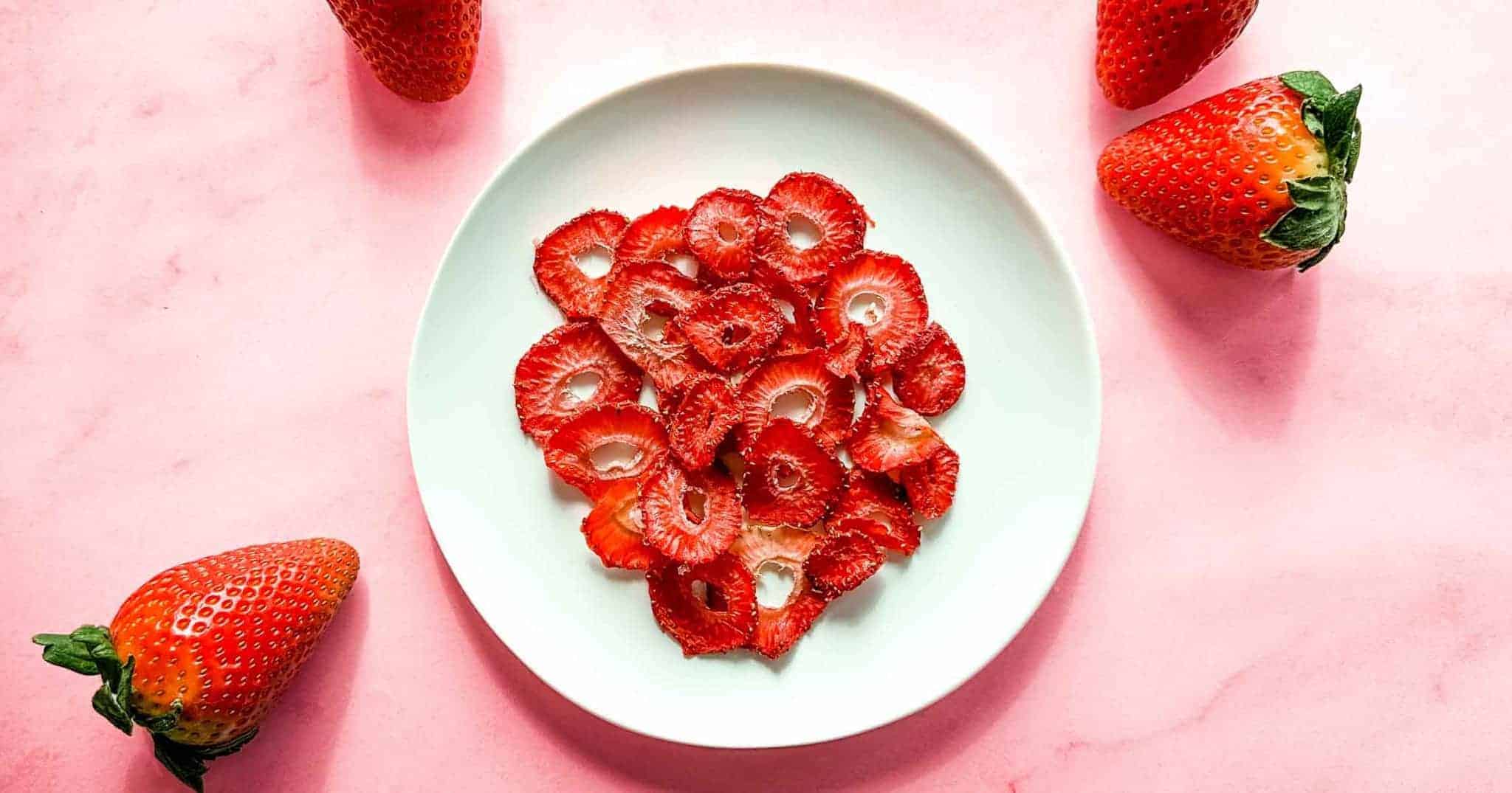 A white plate of air fryer dehydrated strawberries sits on a pink marble background surrounded by fresh strawberries.