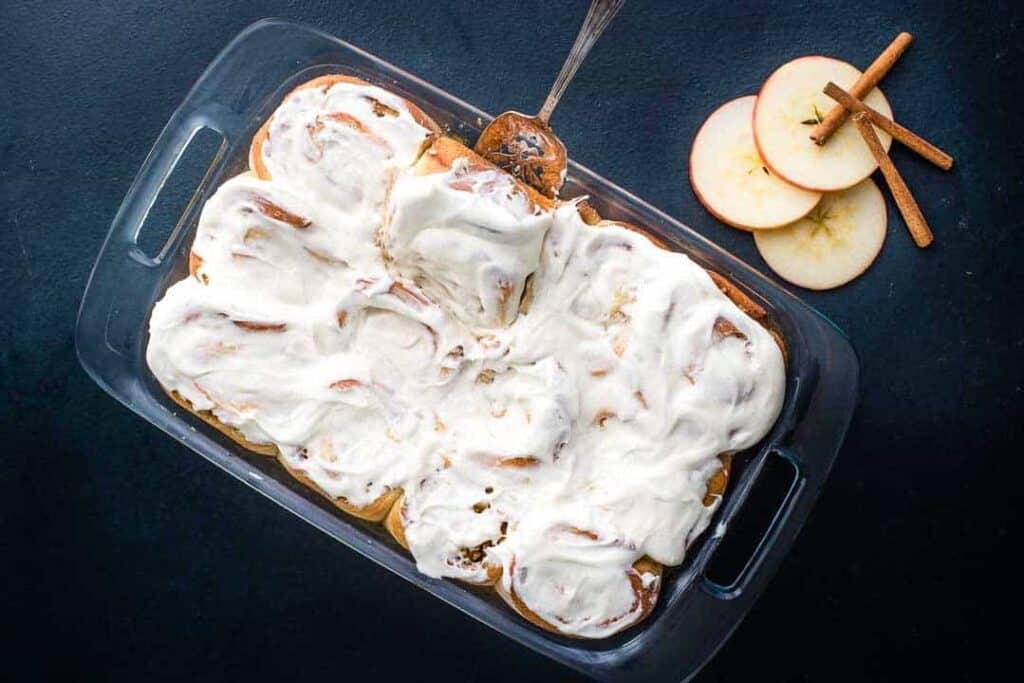 A pan of freshly baked and frosted apple cinnamon rolls on a dark blue background.