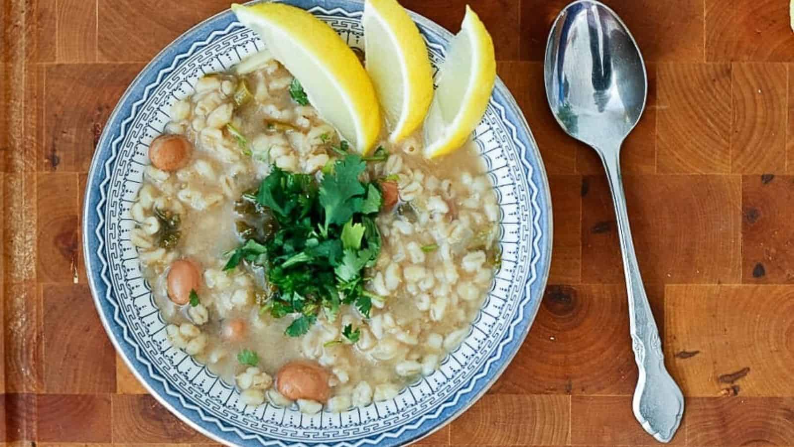 A bowl of barley soup with lemon wedges on a wooden table.