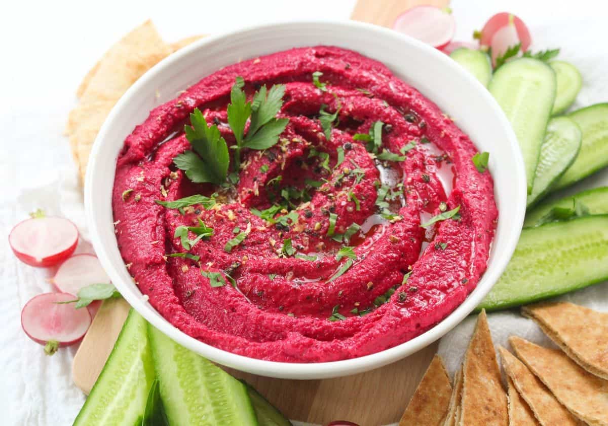 Beet hummus in a white bowl, served with cucumber and pita.