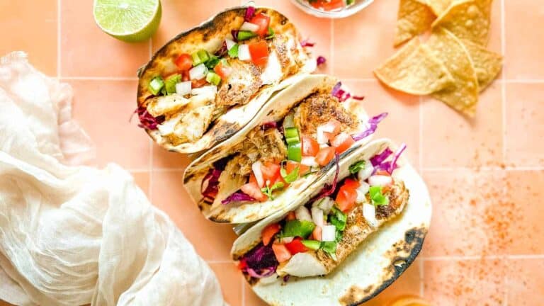 Three fish tacos on a table with limes and tortillas.