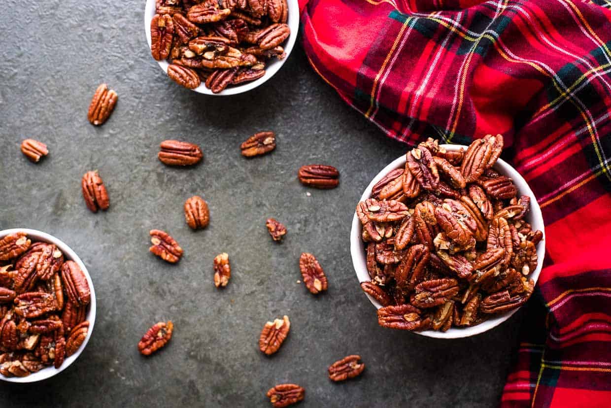 Three bowls of brown butter roasted pecans on a gray background next to a red tartan cloth.
