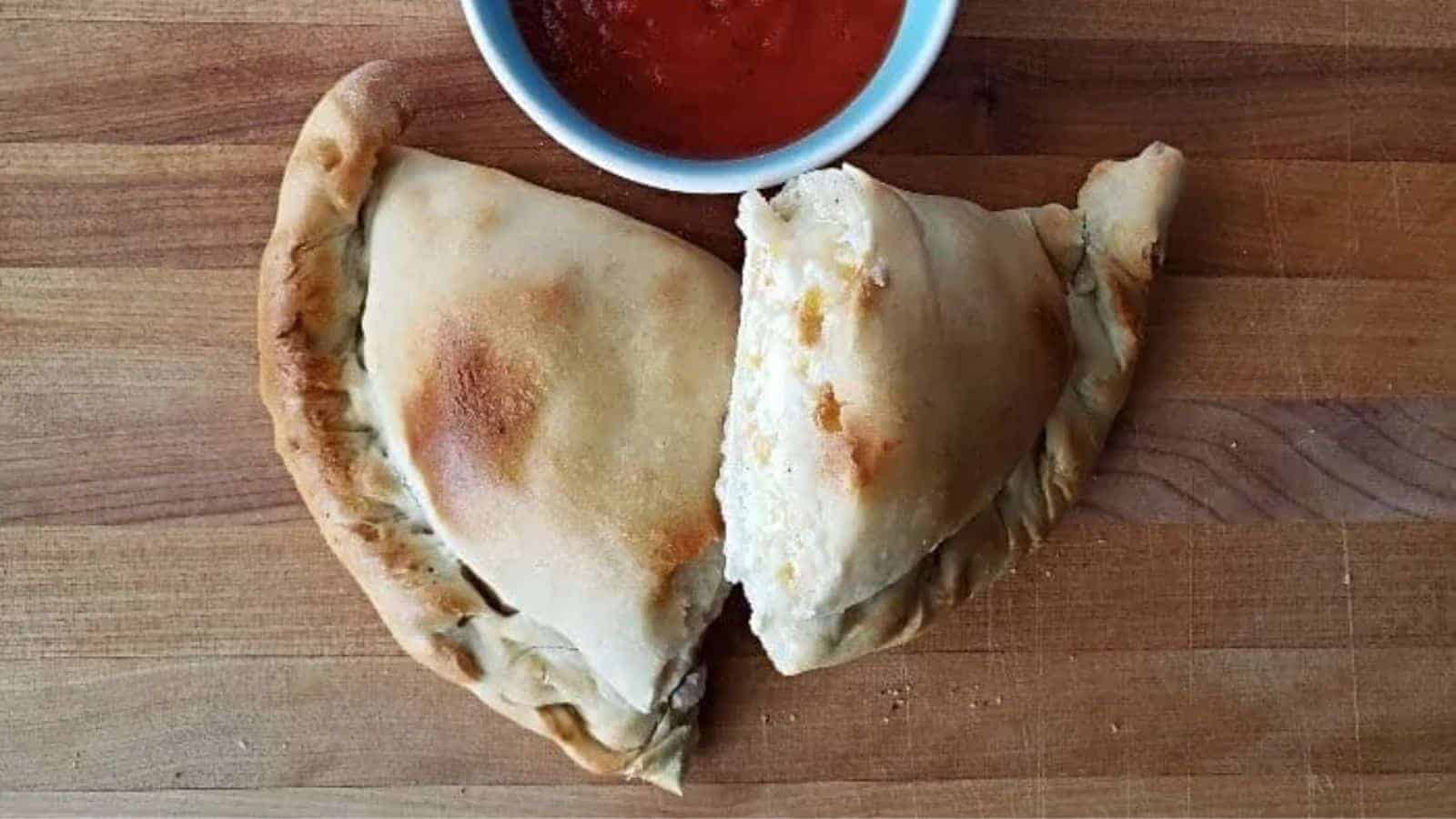 Image shows an overhead shot of a Calzone cut in half on a wooden board with a small bowl of marinara above it.