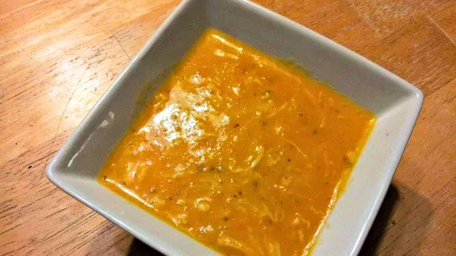 Image shows A bowl of carrot chicken soup on a wooden table.