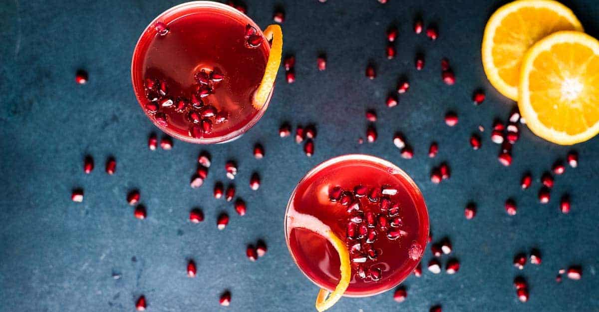 Two champagne pomegranate cocktails in coupe glasses on a dark blue background scattered with pomegranate seeds.