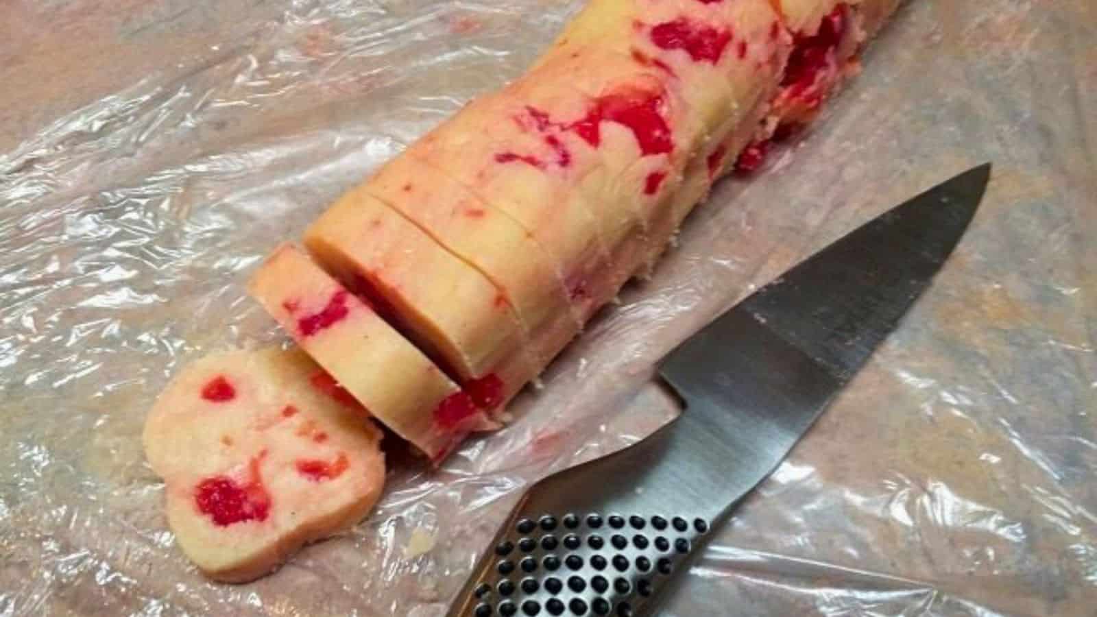 Image shows a knife sitting next to a partially sliced roll of cherry sugar cookies.