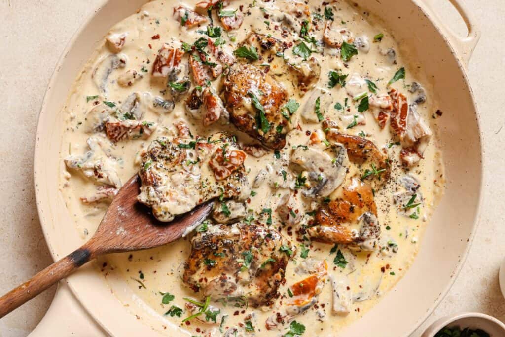 A skillet of chicken with creamy mushroom sauce with a wooden spoon.