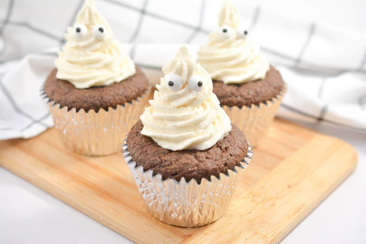 Three chocolate cupcakes with white frosting on a wooden cutting board.