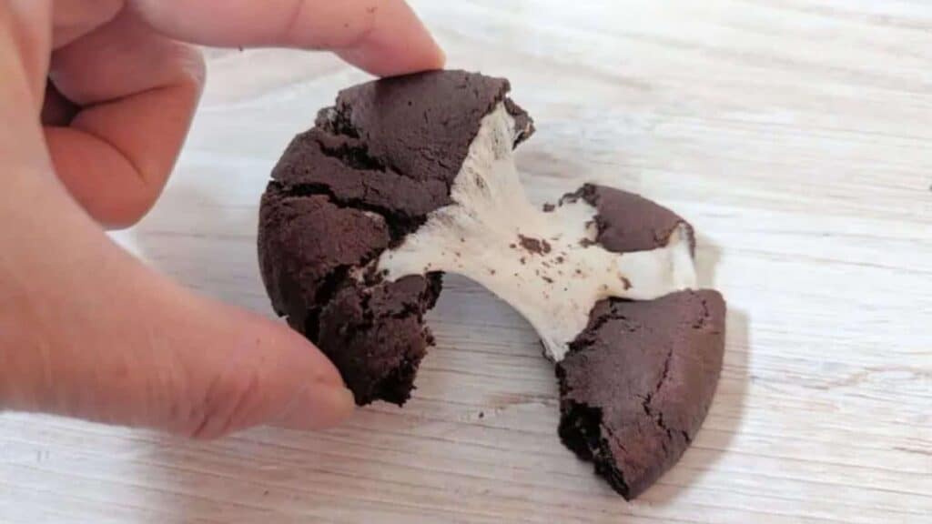 Image shows a Chocolate Marshmallow Cookie being pulled apart on a wooden table.