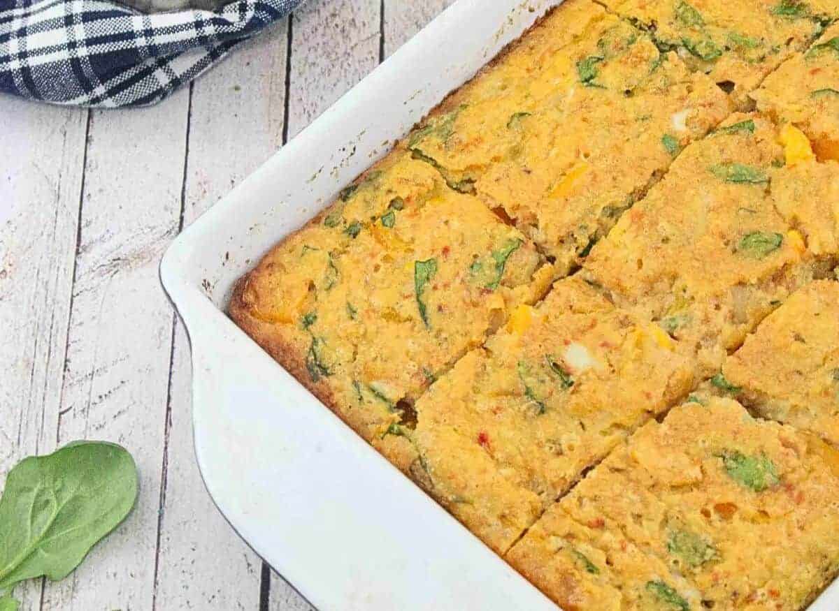 Breakfast casserole on a white plate with more casserole in a pan behind it and a blue linen with steel bowls on top beside it and wooden spoon in front of it.