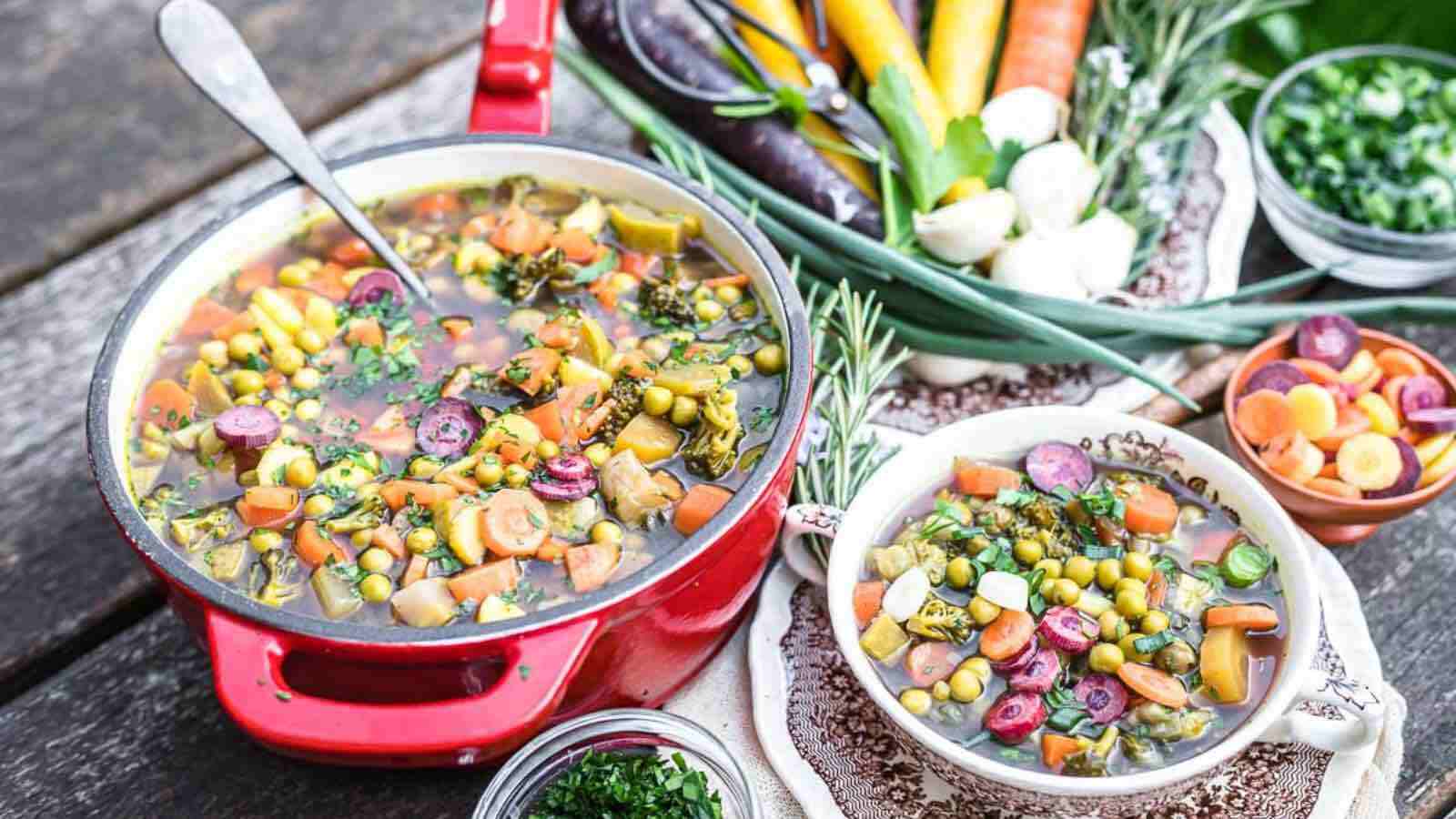 A pot of soup with vegetables and herbs on a wooden table.