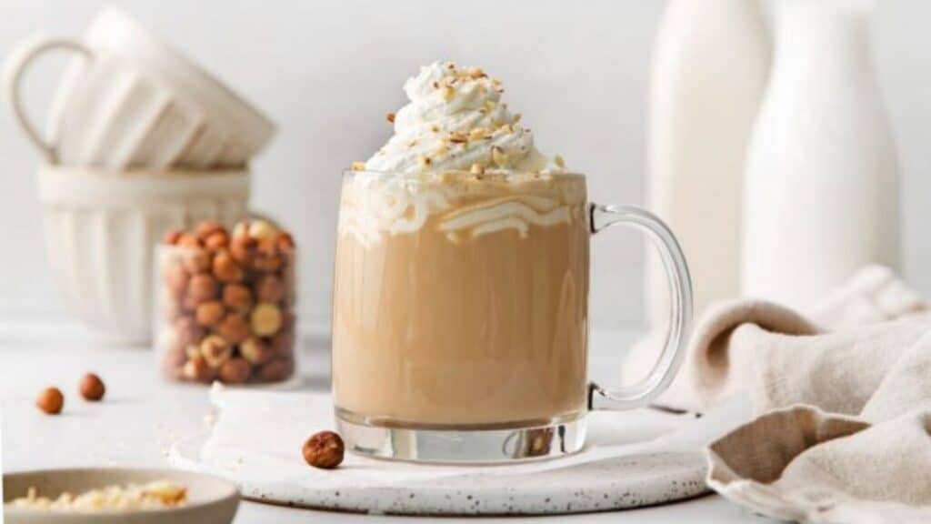 Clear mug with hazelnut latte, whipped cream on top and hazelnuts in the background.