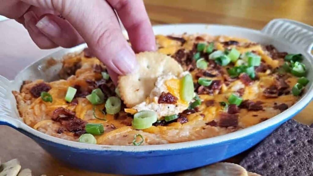 Image shows a hand dipping a cracker into Hot Bacon Cheddar Dip in a ceramic dish.