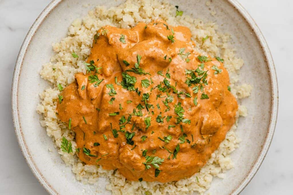 A bowl of butter chicken with cauliflower rice on a white plate.