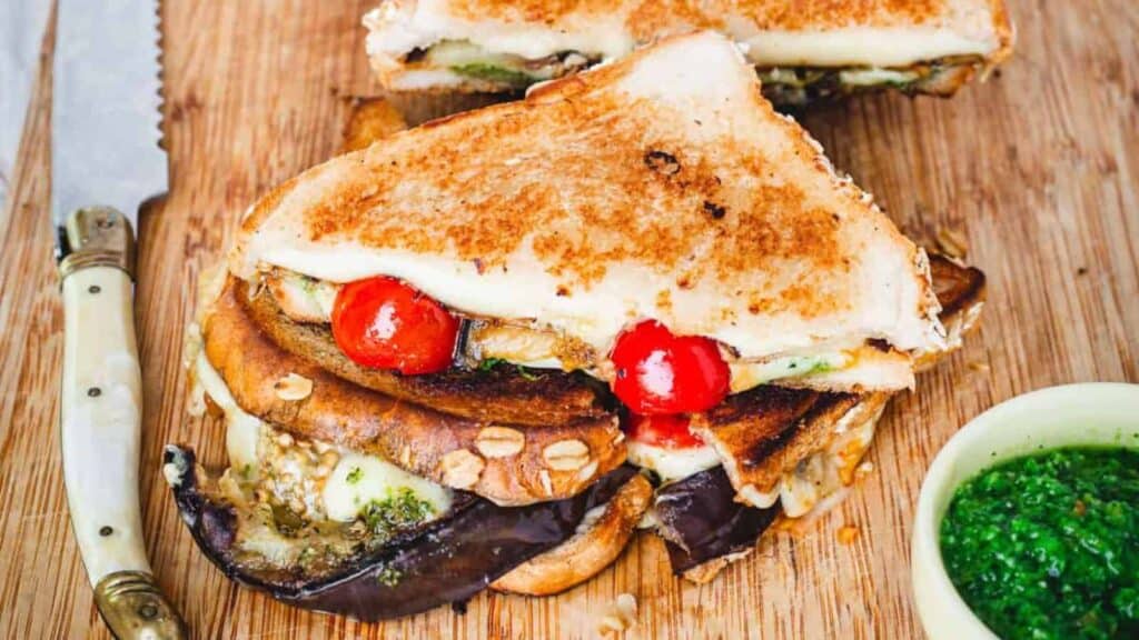 A grilled sandwich with mushrooms, tomatoes and pesto on a cutting board.